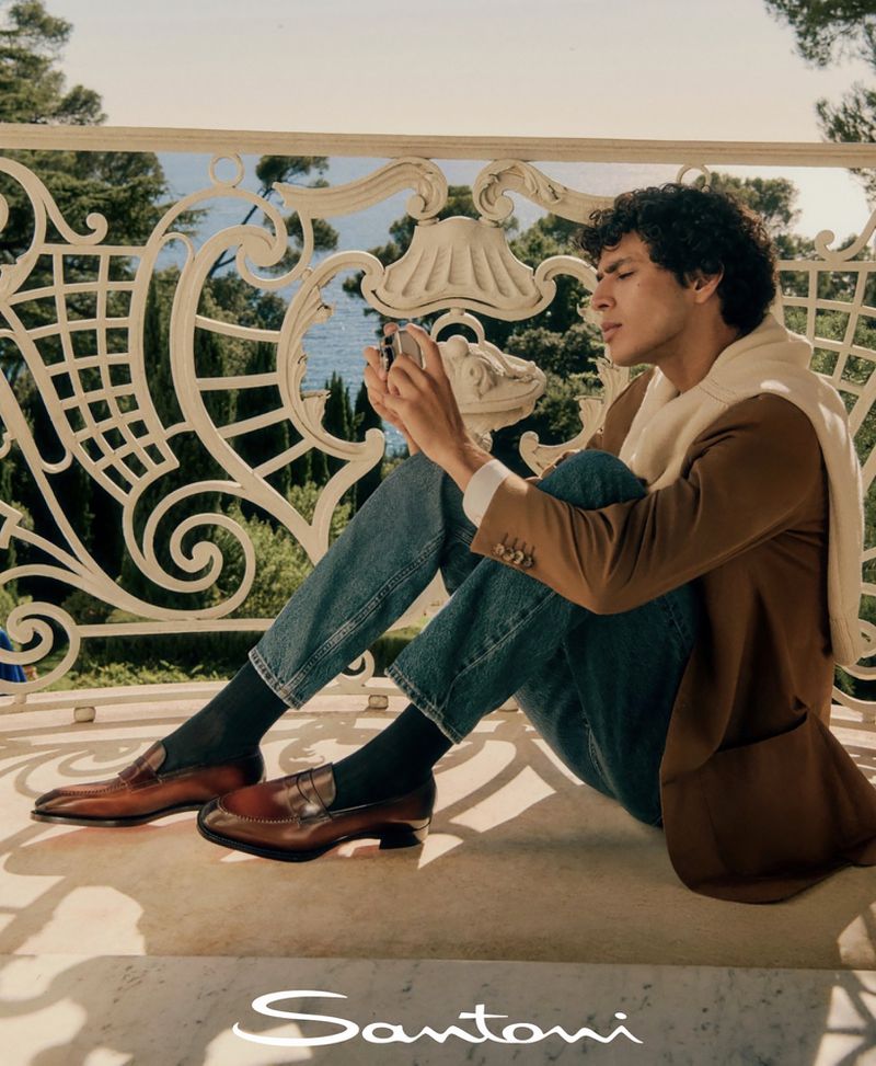 A young man sits on a balcony overlooking the sea while looking at his phone, wearing a brown jacket and jeans with leather loafers.
