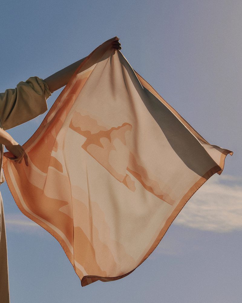 Person holds a peach colored silk scarf against a bright blue sky. The smooth fabric is translucent and flowing in the wind.