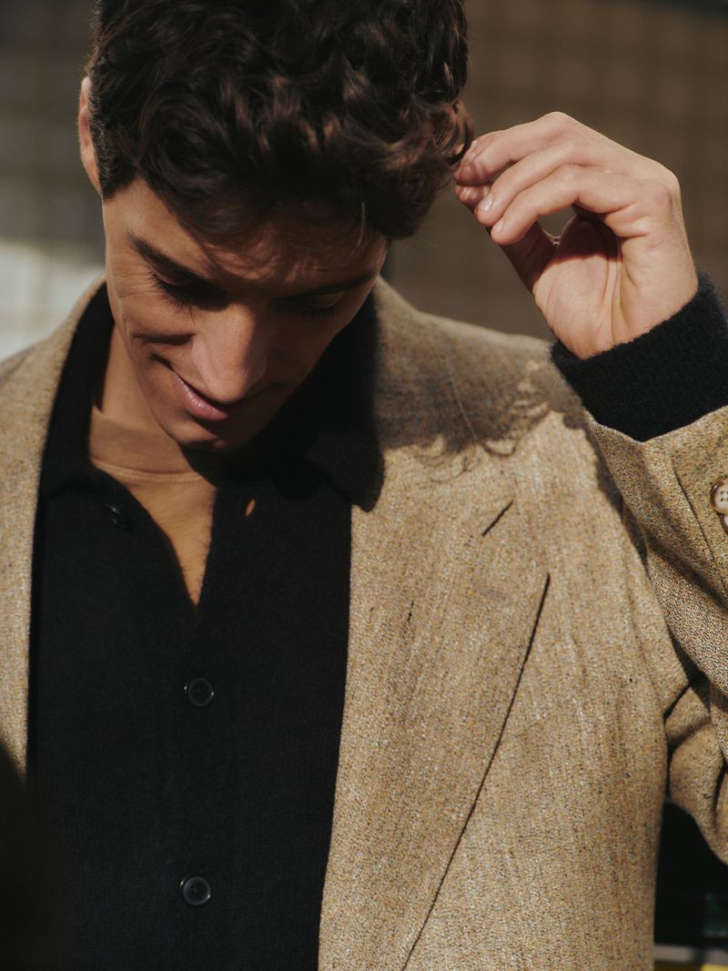 Man in a tan blazer and black shirt touches his hair in a close-up studio shot, with soft lighting enhancing the fabric textures.