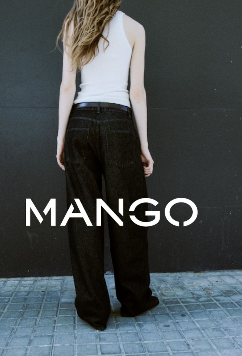Fashionable woman in a white tank top and black jeans strikes a pose against a grey wall and concrete floor, showcasing the Mango brand logo.