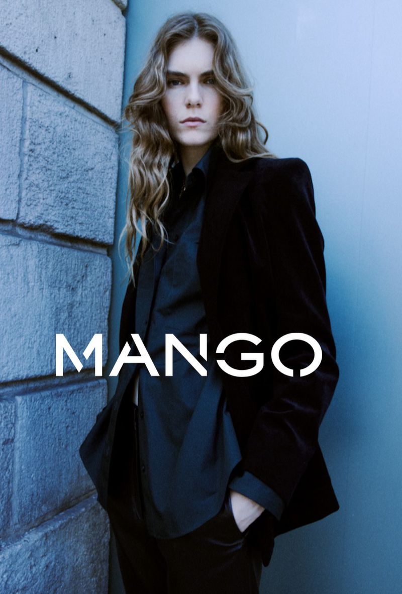 Fashion portrait of a young adult female with long wavy hair, wearing a black velvet blazer and trousers, with the MANGO logo.