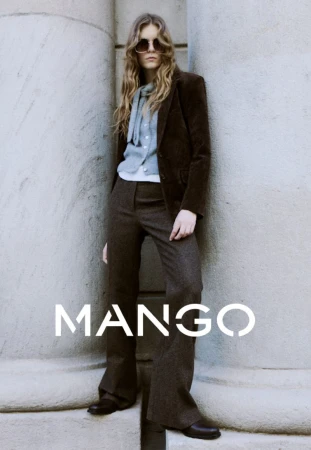 A young woman poses in a brown corduroy blazer, grey trousers, and sunglasses against a textured, neutral wall for a fashion advertisement.