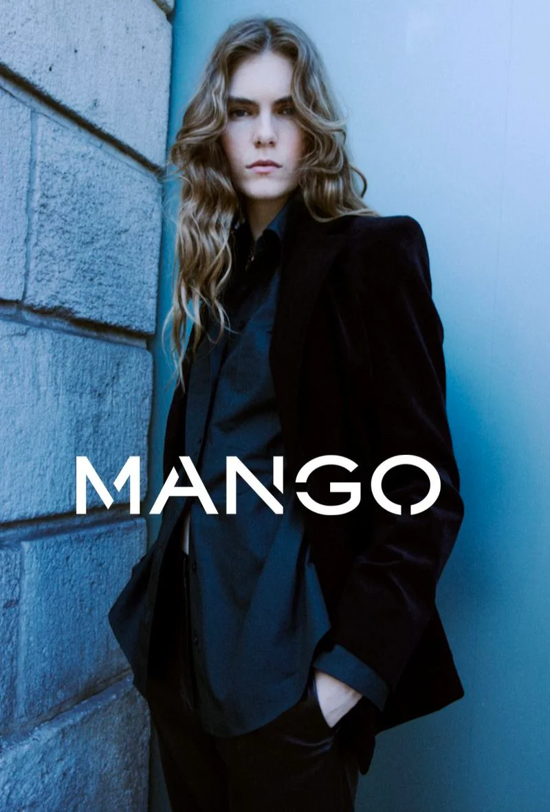 Model with long wavy hair wearing a black blazer and shirt against a stone wall and blue background with Mango text overlay