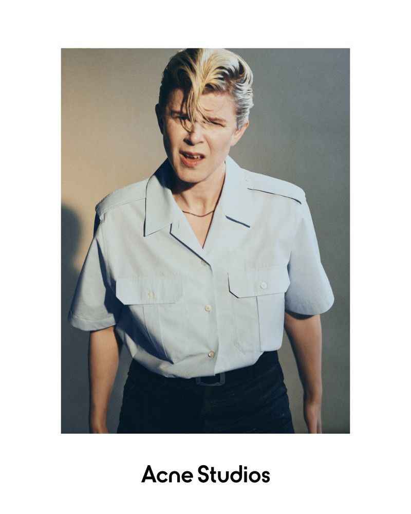 Portrait of a blonde adult woman in a light blue short-sleeved shirt and dark pants against a neutral background indoors.