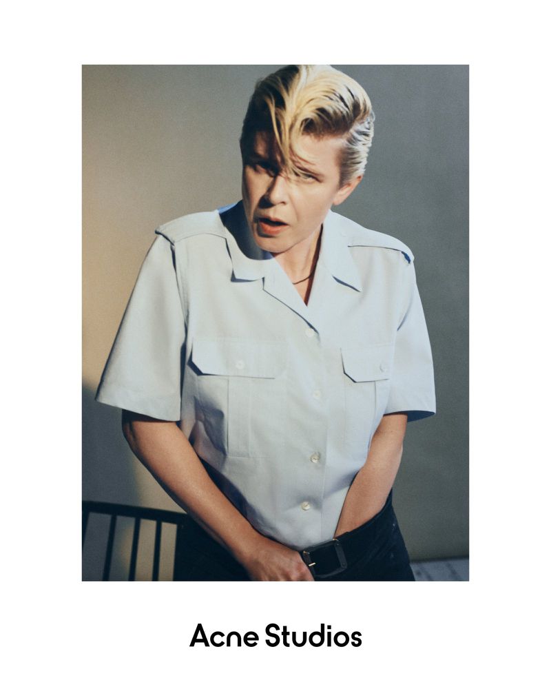 Stylish portrait of an adult woman with a blonde, swept-back hairstyle, wearing a blue shirt and black pants indoors.