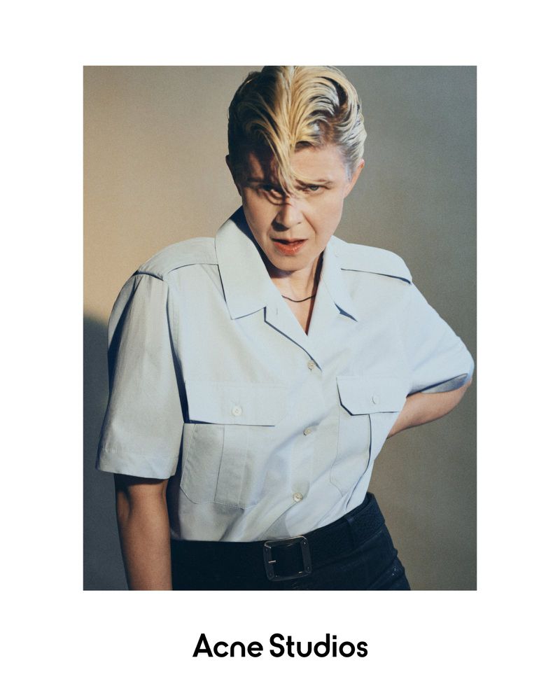 Portrait of a blonde-haired woman in a light blue button-down shirt and black pants, captured indoors against a neutral backdrop.