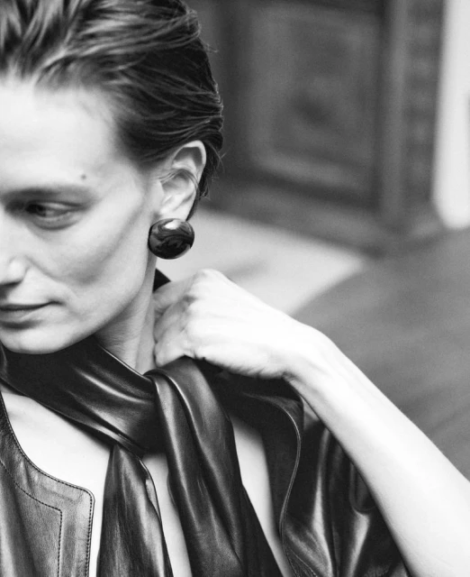 Black and white portrait of a woman with slicked-back hair wearing a black jacket, scarf, and round earrings, with a blurred background.