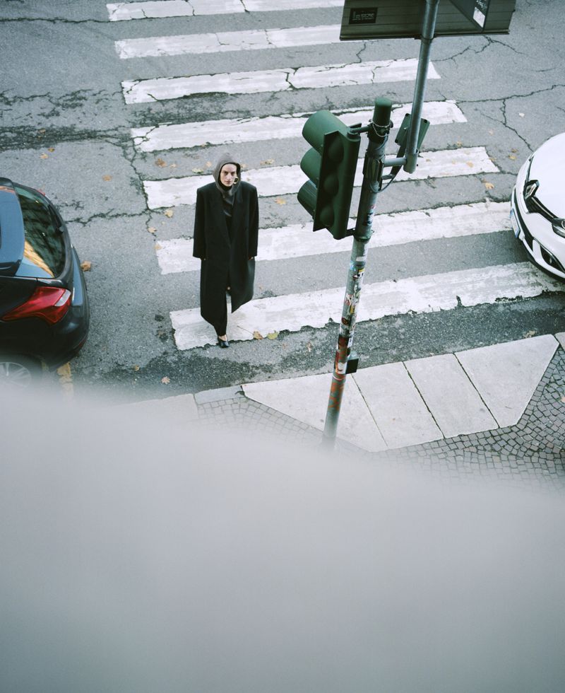 Woman crosses cracked crosswalk, wearing dark coat and hood; traffic light and cars present, shot from a high angle in an urban setting.