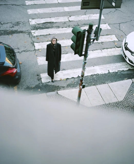 Woman crosses cracked crosswalk, wearing dark coat and hood; traffic light and cars present, shot from a high angle in an urban setting.
