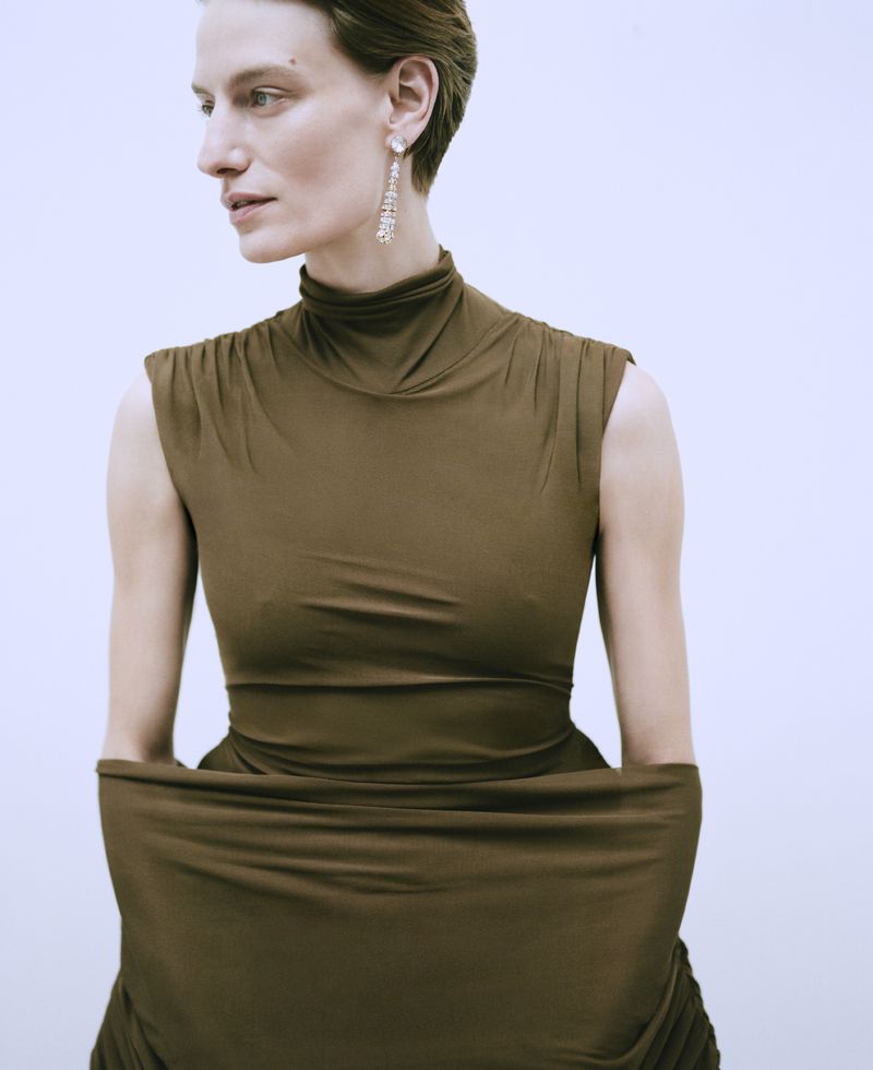 Woman in a draped brown dress with matching sleeves and chandelier earrings poses against a plain white backdrop in a studio setting.