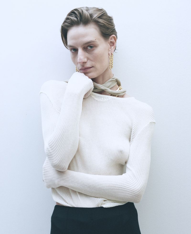 Indoor portrait of a slender adult woman with short hair wearing a beige top, black pants and statement gold earrings against a white wall.
