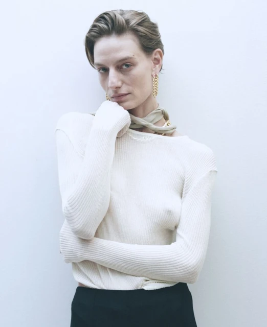 Indoor portrait of a slender adult woman with short hair wearing a beige top, black pants and statement gold earrings against a white wall.