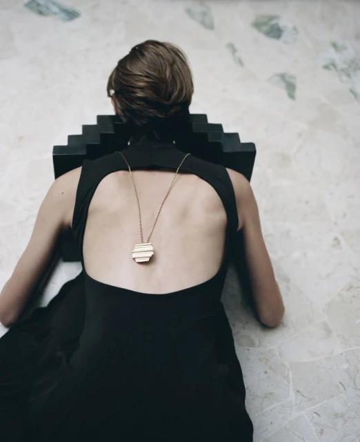 Back view of a woman in a black dress with a geometric pendant necklace, kneeling indoors on a patterned marble floor.