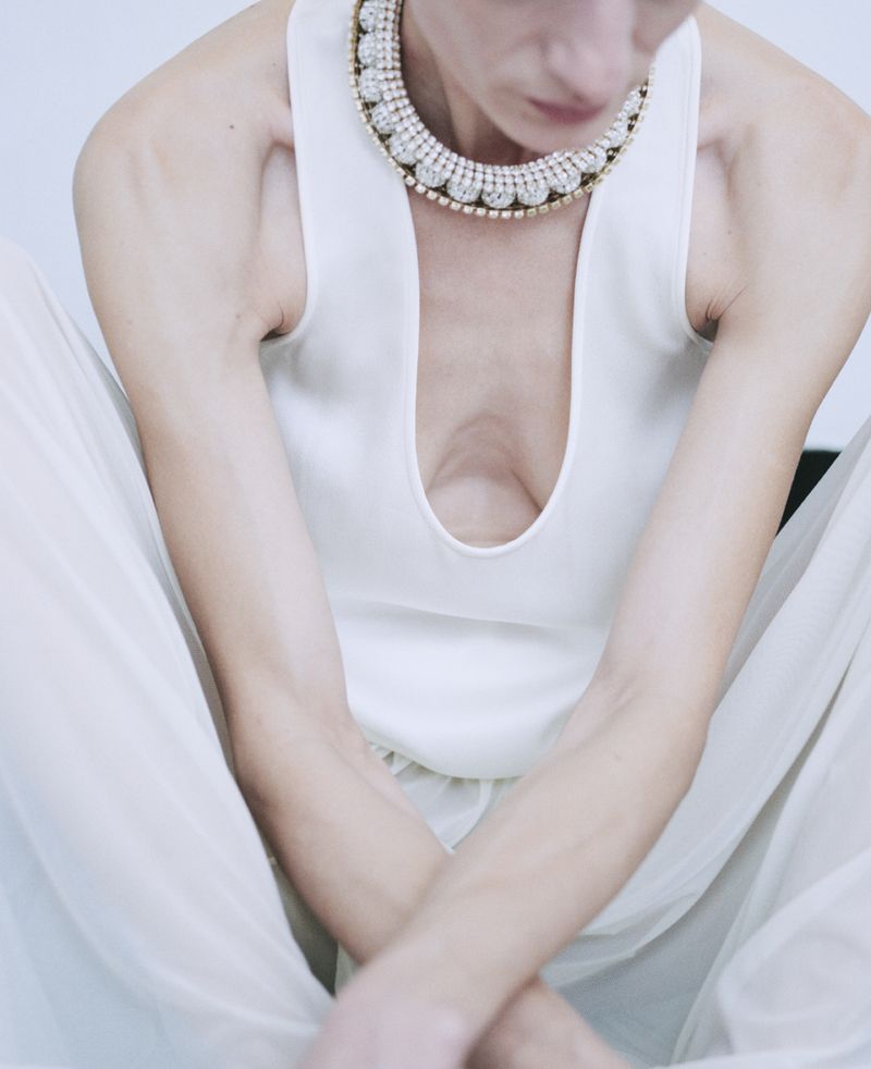Woman in a white tank top and palazzo pants sits with arms crossed, wearing a large, jeweled necklace, in a brightly lit studio setting.