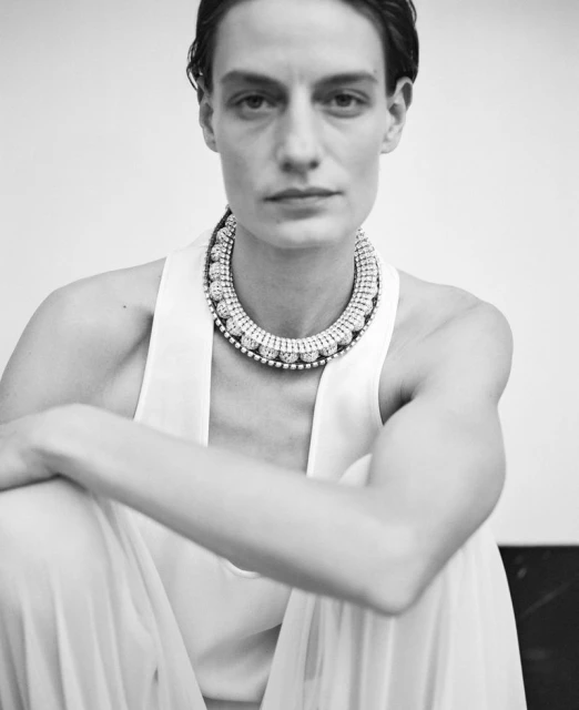 Elegant woman in a sleeveless dress and a statement jeweled necklace poses indoors against a muted wall in a black-and-white portrait.