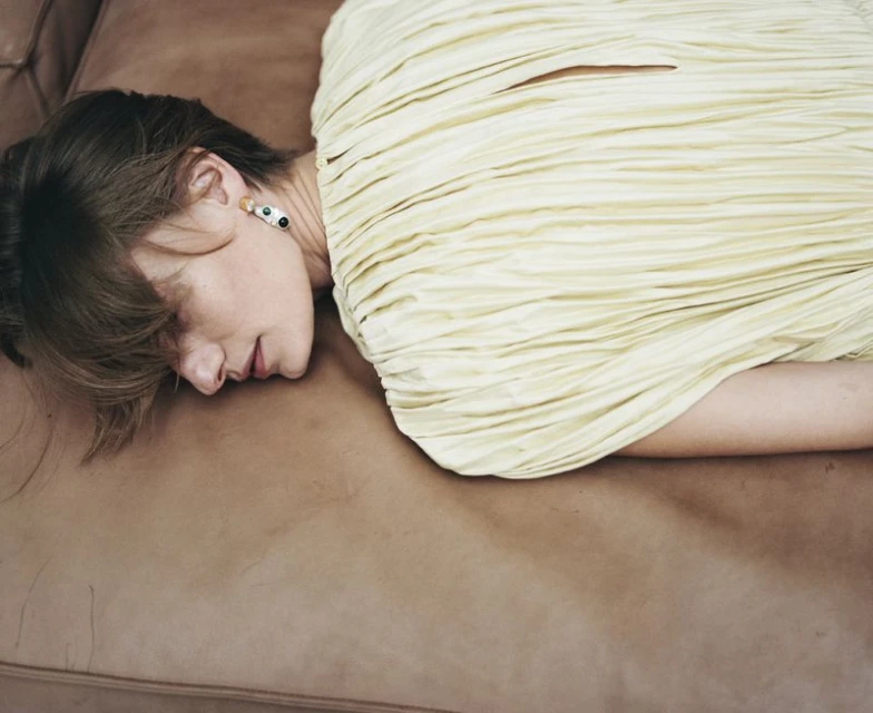 Adult female lying on a brown leather couch, wearing a pleated light yellow dress and colourful circular earrings. Studio lighting.