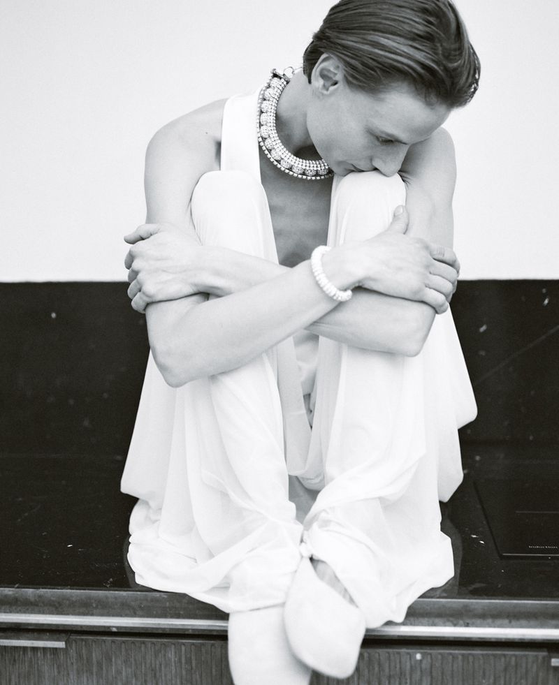Monochrome portrait of a woman in a white dress and pearl jewelry sitting with her legs crossed, embracing her knees.