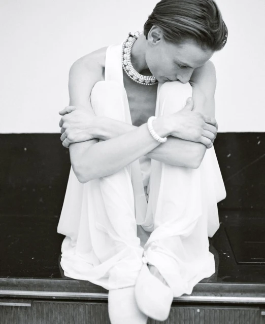 Monochrome portrait of a woman in a white dress and pearl jewelry sitting with her legs crossed, embracing her knees.