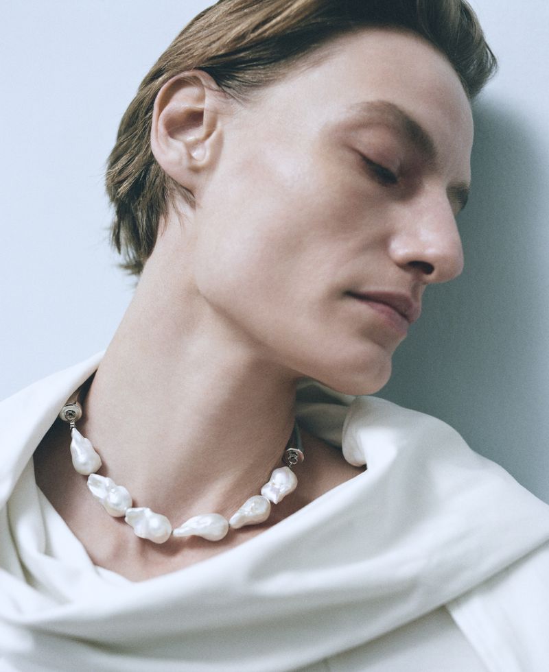 Close-up of a person wearing a baroque pearl necklace, styled with a white draped top against a light blue studio backdrop.