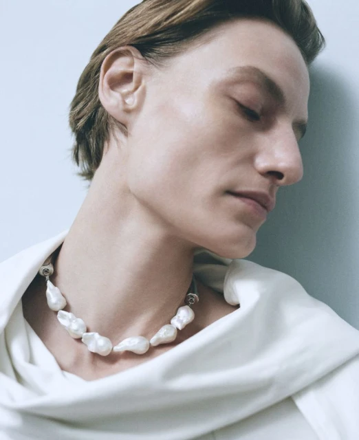 Close-up of a person wearing a baroque pearl necklace, styled with a white draped top against a light blue studio backdrop.