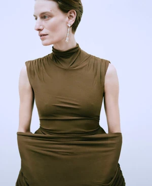 Model wearing a brown sleeveless top with a high collar and long crystal earrings against a white background
