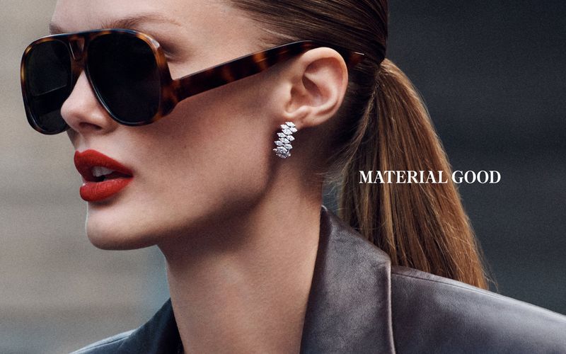 Close-up of a woman with sunglasses and red lipstick wearing diamond earrings, accessorized with a leather jacket against a grey backdrop.