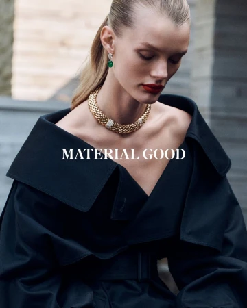 A blonde woman in a black trench coat poses in an outdoor fashion shot, showcasing gold jewelry with diamond accents and green gemstone earrings.