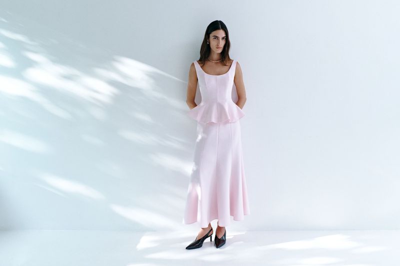 Elegant woman in a light pink gown standing against a white wall with soft light and shadows, accessorized with a necklace and dark shoes.