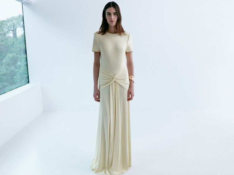 Woman in flowing cream dress stands in bright room near window. Elegant fashion, soft lighting, minimalist style.