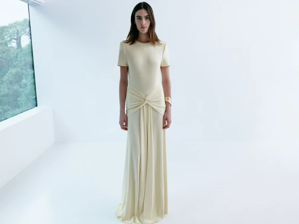 Woman in flowing cream dress stands in bright room near window. Elegant fashion, soft lighting, minimalist style.