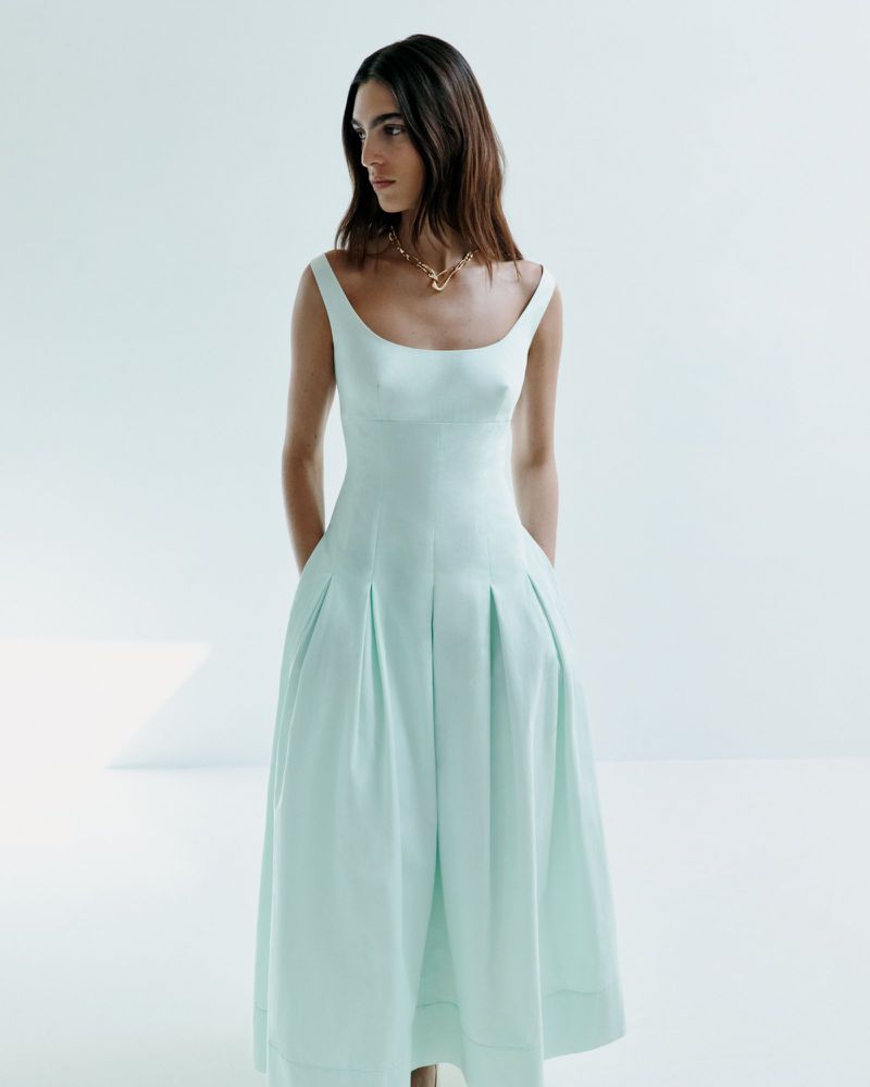 Elegant woman in a light blue dress and gold necklace poses in a bright studio setting with a clean, minimalist aesthetic for fashion.