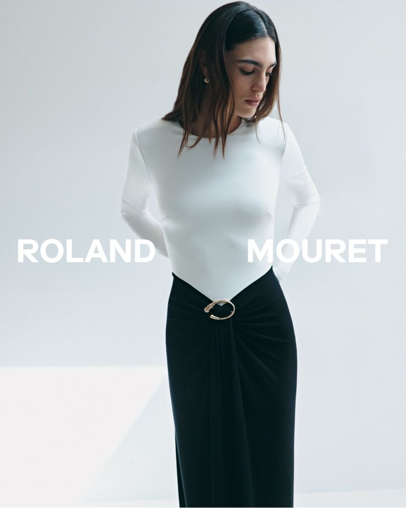 Elegant woman poses in a white long-sleeve shirt and black skirt with an embellishment against a clean white backdrop in a studio shot.