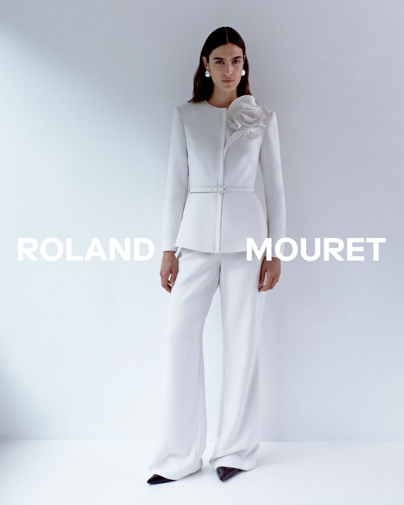 Fashion shoot featuring a woman wearing a white Roland Mouret blazer with a flower detail and wide-leg pants, posing in a studio.