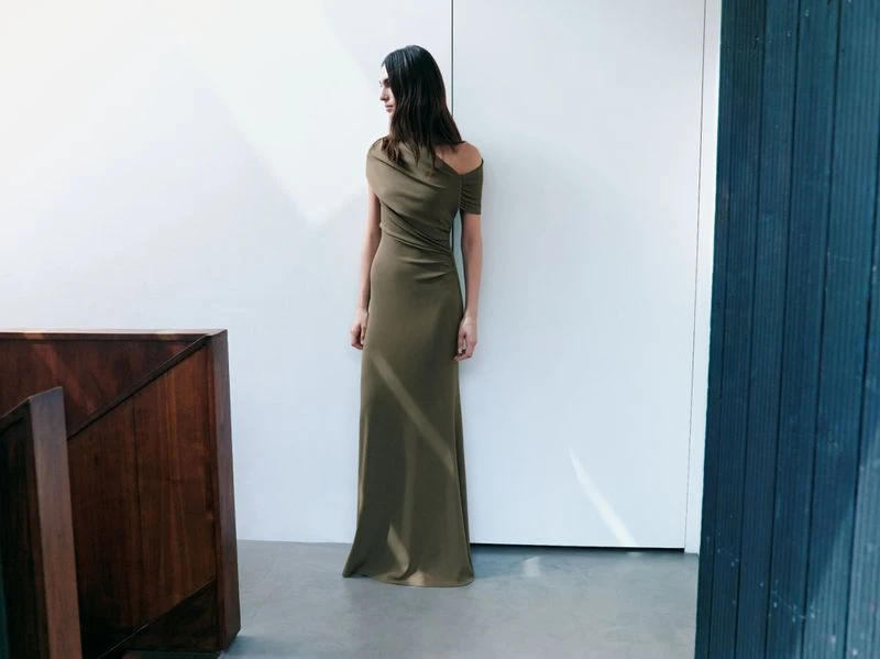 Elegant woman in an olive-green draped gown stands indoors against a white wall, showcasing contemporary fashion and minimalist style.