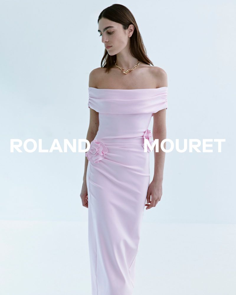 A fashionable woman in a light pink off-the-shoulder evening gown poses in a bright, minimalist setting, accessorized with a gold necklace.