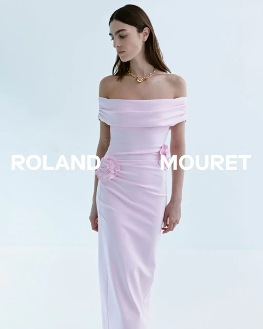 A fashionable woman in a light pink off-the-shoulder evening gown poses in a bright, minimalist setting, accessorized with a gold necklace.