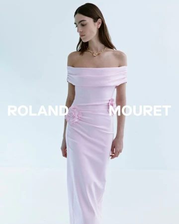 A fashionable woman in a light pink off-the-shoulder evening gown poses in a bright, minimalist setting, accessorized with a gold necklace.