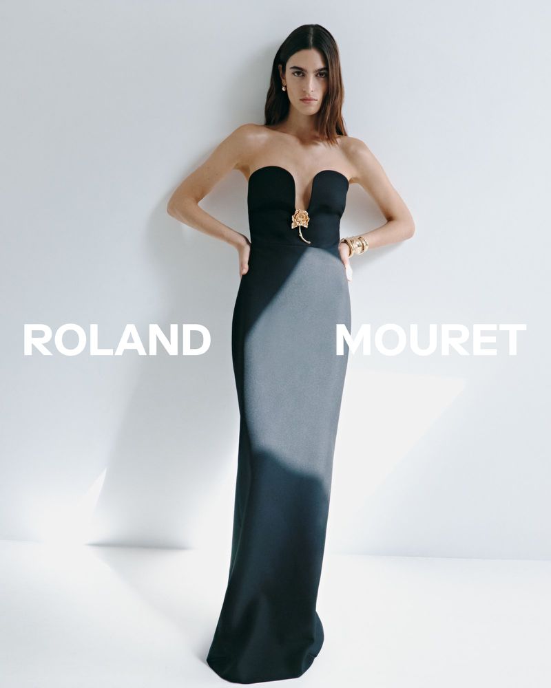 Elegant woman in a Roland Mouret gown poses against a white backdrop, accessorized with gold jewelry, showcasing a sophisticated and modern fashion aesthetic.