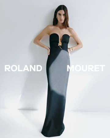 Elegant woman in a Roland Mouret gown poses against a white backdrop, accessorized with gold jewelry, showcasing a sophisticated and modern fashion aesthetic.
