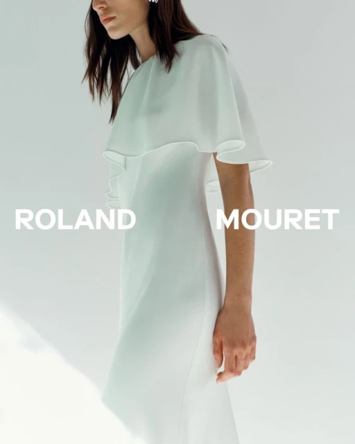 A model wears a white Roland Mouret dress with a flowing cape against a light background in a fashion advertisement.