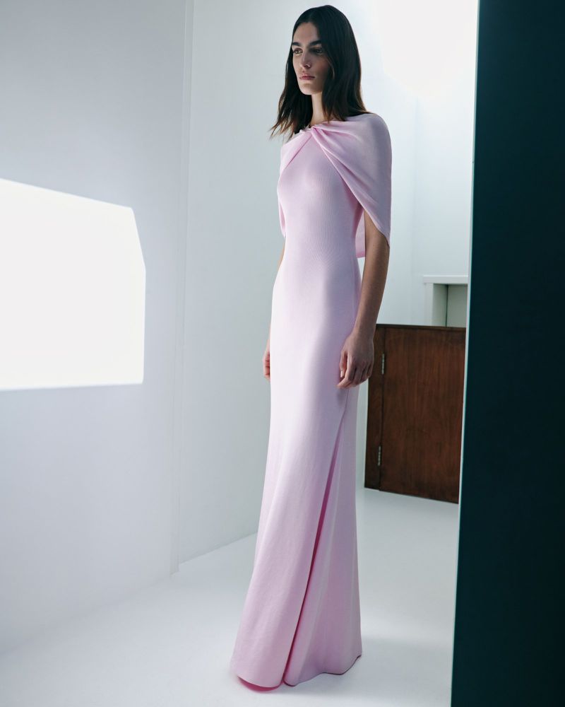 Elegant woman in a light pink gown, posing in a white room with soft lighting for a fashion editorial, capturing the dress's graceful design.