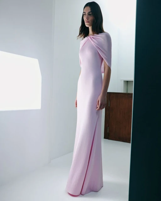 Elegant woman in a light pink gown, posing in a white room with soft lighting for a fashion editorial, capturing the dress's graceful design.