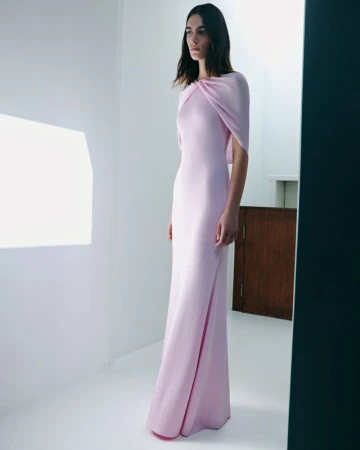 Elegant woman in a light pink gown, posing in a white room with soft lighting for a fashion editorial, capturing the dress's graceful design.