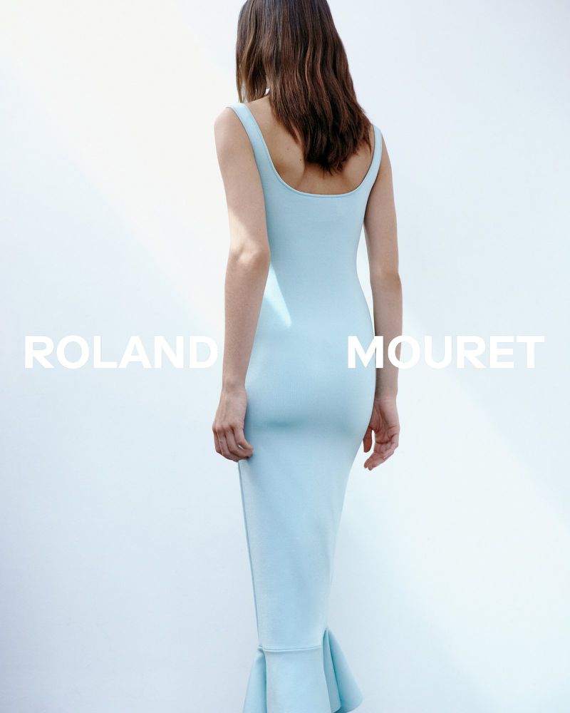 Woman in a fitted light blue dress stands against a bright white background, back to camera, minimalist fashion photography.