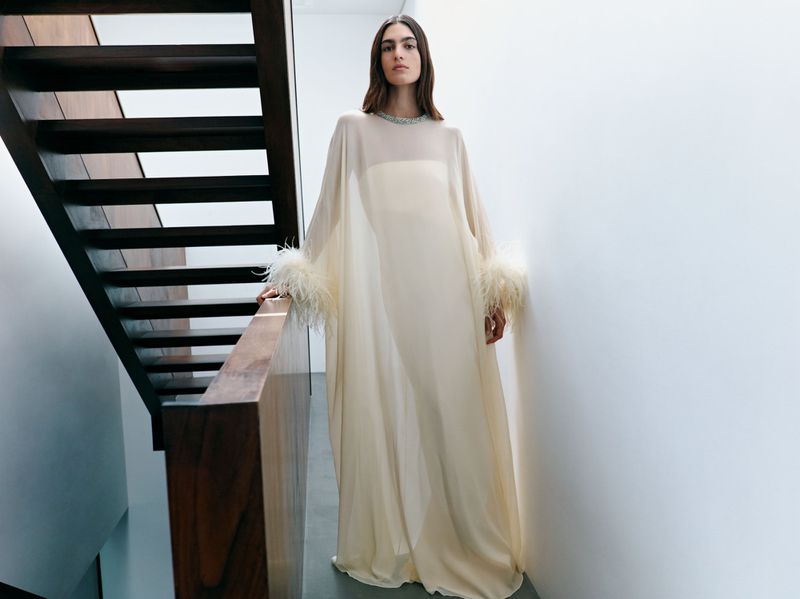 Fashion shot of a woman in a flowing, off-white gown with feathered sleeves, posing on a staircase with a modern, minimalist aesthetic.