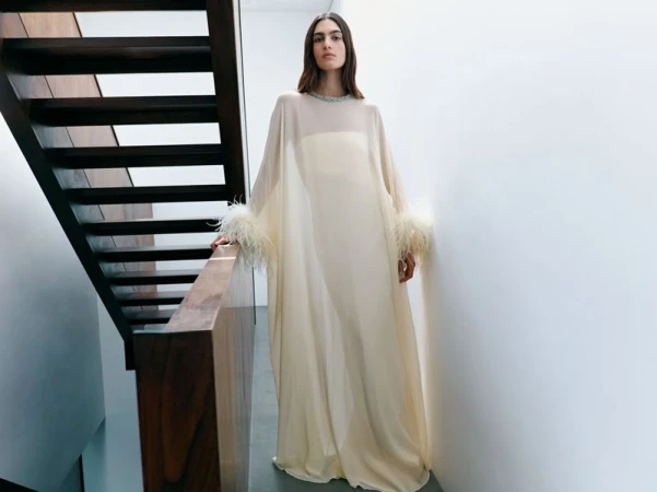 Fashion shot of a woman in a flowing, off-white gown with feathered sleeves, posing on a staircase with a modern, minimalist aesthetic.