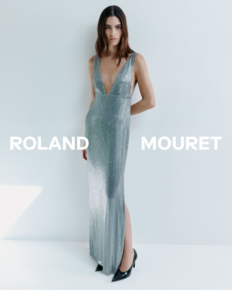 A fashionable adult female posing in a floor-length shimmering silver dress and high heels against a white background for a fashion shot.