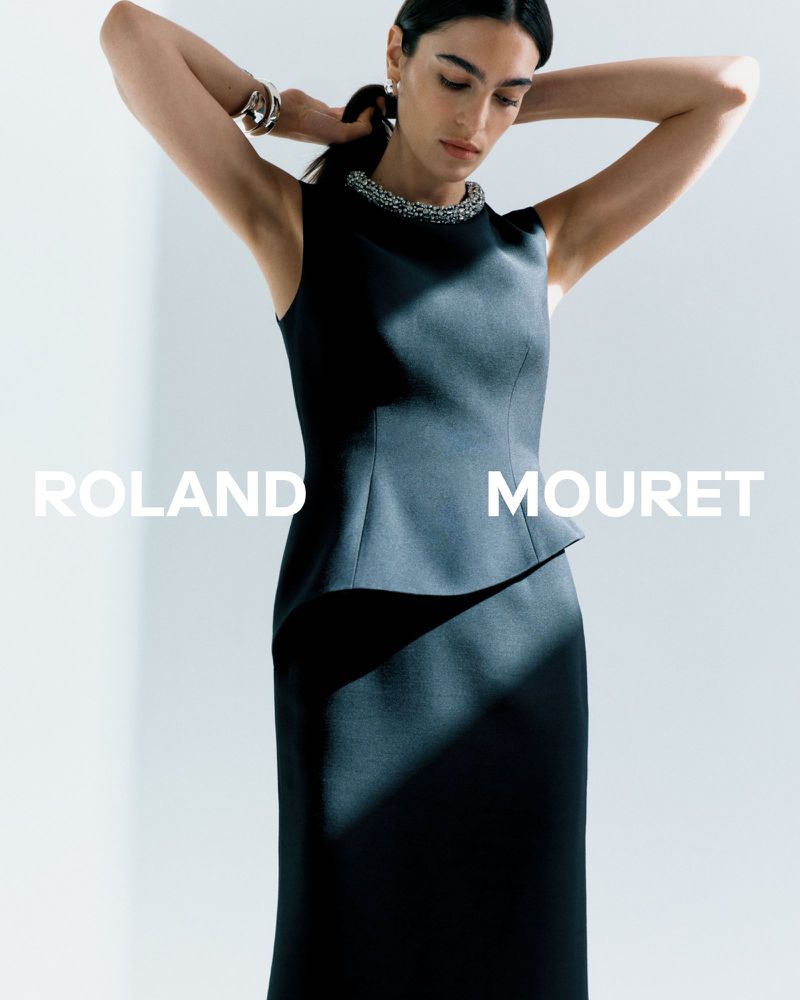Elegant woman in a dark sleeveless dress with embellished neckline, silver bracelets, and hair tied back, poses against a light background.