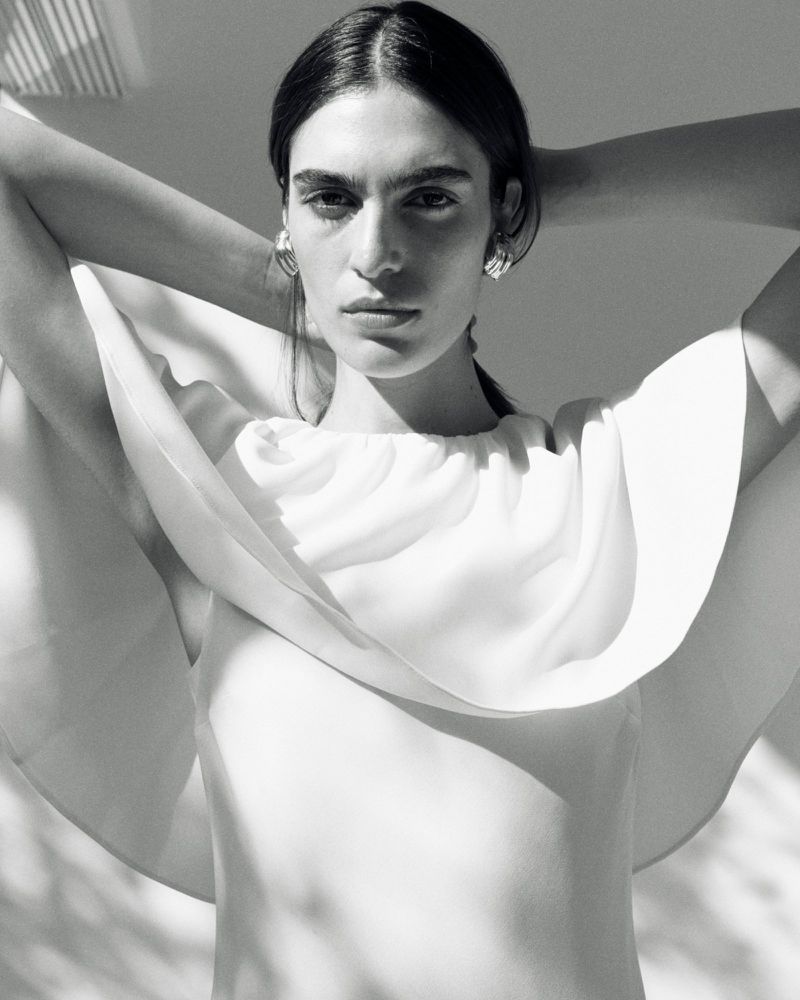 Elegant fashion shot of a woman in a white dress with unique shoulder design, complemented by statement earrings, set against a minimalist backdrop.