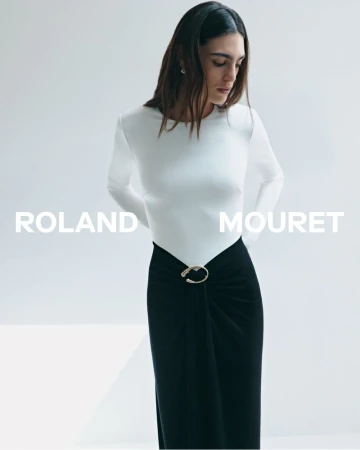 Model wearing a white long-sleeve top and black skirt with a gold belt buckle against a plain white studio background.